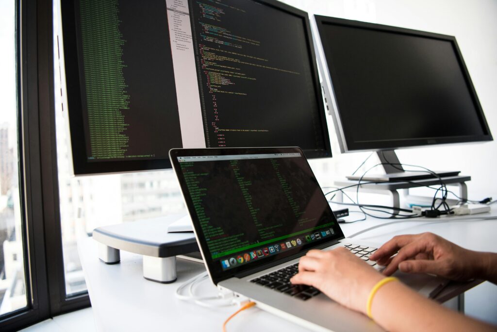 pexels-photo-1181675-1181675 A developer writes code on a laptop in front of multiple monitors in an office setting.